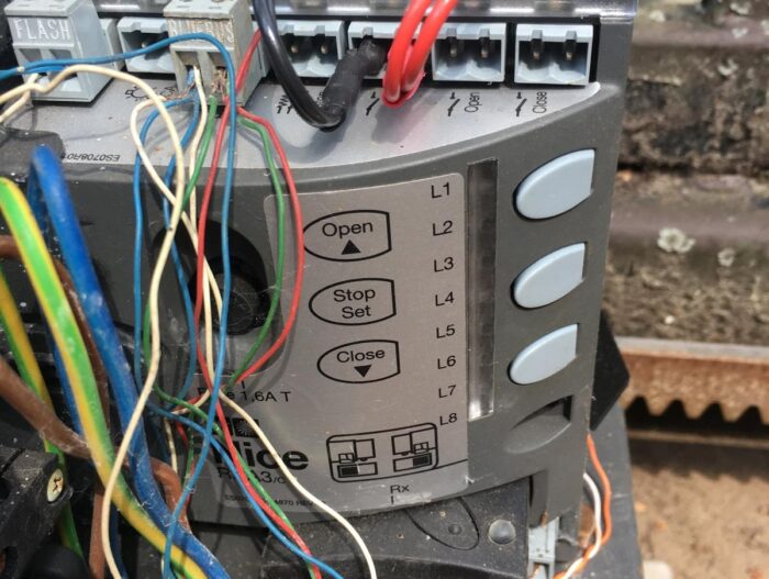 WhatsApp Image 2024-08-04 at 15.39.15 Obraz na stronie internetowej przedstawia szczegółowy panel sterowania używany do naprawy bram, rolet i barier w Warszawie. Panel zawiera wyraźnie oznaczone przyciski „Otwórz”, „Stop/Ustaw” i „Zamknij”. Zawiera również kilka punktów połączeń przewodów oznaczonych jako L1, L2, L3 i L8. W tle znajduje się falista powierzchnia dodająca faktury kompozycji.