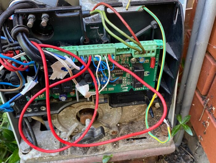 WhatsApp Image 2024-08-04 at 15.40.51 Obraz przedstawia szczegółowy widok otwartego urządzenia mechanicznego z zieloną płytką drukowaną połączoną wieloma kolorowymi przewodami. Ta skomplikowana konstrukcja jest bezpiecznie zamontowana na metalowym uchwycie, otoczona rozrzuconymi liśćmi, co podkreśla środowisko zewnętrzne, zwykle kojarzone z mechanizmami bram i barier w Warszawie.