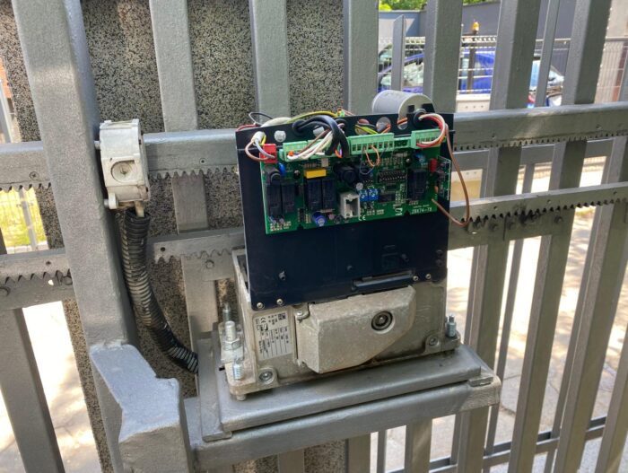 WhatsApp Image 2024-08-04 at 15.51.57 Na zdjęciu widać szczegółowy widok silnika metalowej bramy i jej podzespołów elektronicznych, podkreślając płytkę drukowaną, misternie połączoną z szeregiem przewodów i złączy. Ta konfiguracja jest solidnie przymocowana do ramy bramy. W tle widać scenę uliczną z zaparkowanymi samochodami widocznymi przez pręty bramy, co podkreśla jej miejski charakter w Warszawie.