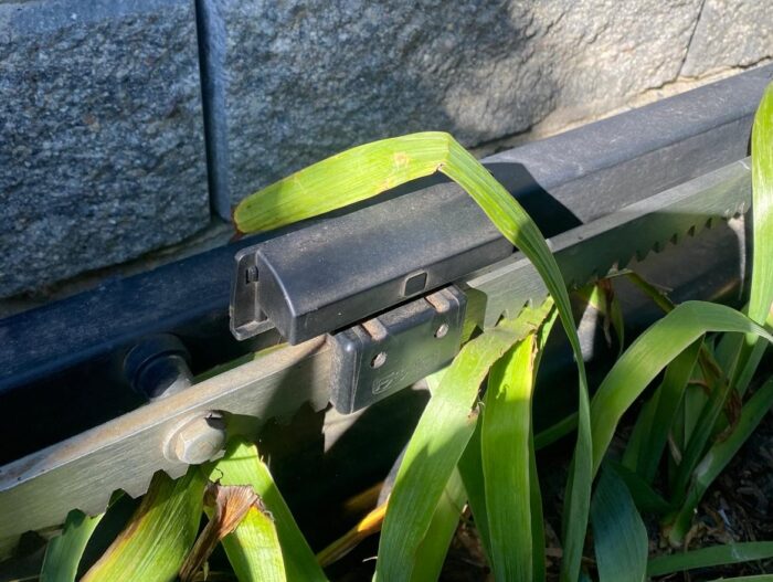 WhatsApp Image 2024-08-04 at 16.00.44 Obraz na naszej stronie internetowej przedstawia szczegółowy widok czarnej metalowej bariery z mechanizmem zębatkowym, umieszczonej pośród bujnej zieleni liści. Umieszczona przy rustykalnym kamiennym murze bariera jest skąpana w świetle słonecznym, tworząc grę światła i cienia na całej scenie.