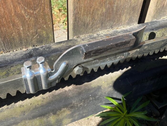 WhatsApp Image 2024-08-04 at 16.00.57 Na dedykowanej warszawskiej stronie internetowej, która specjalizuje się w naprawach bram, rolet i barier, obraz przedstawia szczegółowy widok metalowego mechanizmu przymocowanego do drewnianej bramy. Scena zawiera elementy, takie jak zębata szyna i zasuwka na tle światła słonecznego, które podkreśla ich metaliczny połysk na zniszczonym drewnie. Poniżej cień rzucany przez bramę pada na ziemię, podkreślając przebijającą się przez nią małą zieloną roślinę.