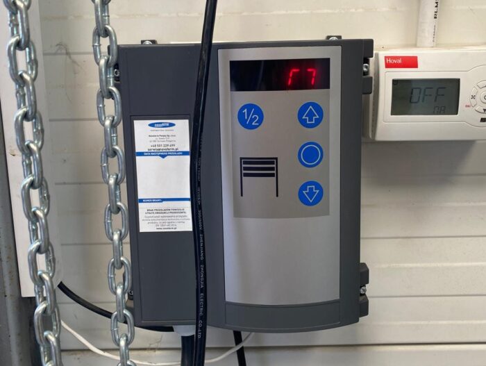 WhatsApp Image 2024-08-10 at 16.26.54 Obraz na stronie internetowej poświęconej naprawie bram, rolet i barier w Warszawie przedstawia panel sterowania przymocowany do ściany. Na wyświetlaczu panelu widnieje „17” i znajduje się kilka przycisków do modyfikowania ustawień. Jest on również połączony z otaczającym go łańcuchem. Obok tego zestawu znajduje się kompaktowe urządzenie wskazujące „WYŁ.” i wyświetlające liczbę „18”.