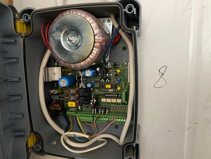 WhatsApp Image 2024-08-10 at 16.29.12 Obraz na stronie internetowej przedstawia otwartą obudowę elektryczną, odsłaniającą płytkę drukowaną wypełnioną elementami, takimi jak kondensatory, rezystory i transformator połączony różnymi przewodami. Obudowa jest przymocowana do jasnego tła, gdzie w pobliżu widoczna jest mała, ręcznie narysowana cyfra „8”. Ten wizualny szczegół jest zgodny z tym, że strona koncentruje się na naprawach bram, rolet i barier w Warszawie.