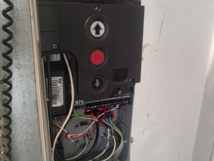 WhatsApp Image 2024-08-10 at 16.49.55 Obraz na stronie internetowej przedstawia szczegółowy widok otwartego panelu sterowania elektrycznego, zaprojektowanego specjalnie dla usług naprawy bram, rolet i barier w Warszawie. Wewnątrz zauważysz szereg odsłoniętych przewodów w wielu kolorach, splecionych wokół płytki drukowanej. Najbardziej widoczne są przyciski ze strzałkami w górę i w dół obok czerwonego przycisku alarmowego. Po lewej stronie panelu widoczny jest solidny łańcuch na tle uporządkowanego tła.