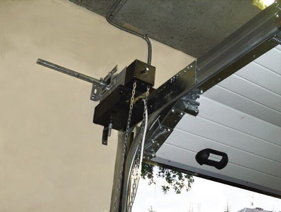 avtomatika-dlya-sektsionnyh-vorot-svoimi-rukami-1 Obraz na stronie internetowej przedstawia garaż w Warszawie z częściowo otwartymi drzwiami wyposażonymi w otwieracz montowany na suficie. Mechanizm obejmuje solidne metalowe elementy i system łańcuchowy. Na panelu drzwi wyraźnie widać czarny uchwyt, co zwiększa ich funkcjonalność. Obok tej konfiguracji, okablowanie elektryczne jest starannie ułożone, podkreślając techniczne aspekty usług naprawy i instalacji bram oferowanych przez witrynę.