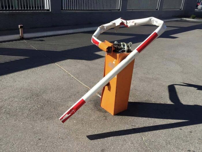 Obraz na stronie internetowej przedstawia uszkodzoną barierę parkingową z czerwonymi i białymi pasami. Jest ona podłączona do pomarańczowej skrzynki sterowniczej i umieszczona na utwardzonej powierzchni, gdzie rzuca wyraźne cienie.