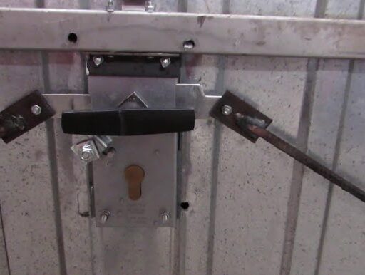 hq720 Na zdjęciu widać szczegółowy widok mechanizmu zamka na metalowych drzwiach garażowych. Dwa solidne metalowe pręty rozciągają się poziomo, a elegancki czarny uchwyt i otwór na klucz zapewniają bezpieczeństwo. Cały zestaw jest zamontowany na polerowanej ocynkowanej powierzchni stalowej, ilustrując wysokiej jakości materiały używane do napraw i instalacji bram w Warszawie.