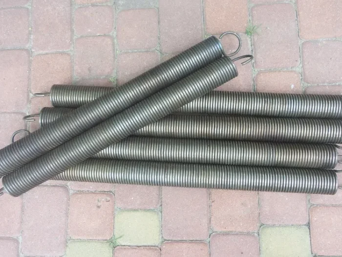image (1) Obraz na stronie internetowej poświęconej naprawom bram, rolet i barier w Warszawie przedstawia zbiór pięciu dużych cylindrycznych metalowych sprężyn, każda wyposażona w haki na obu końcach. Te sprężyny spiralne są wyświetlane na tle kwadratowych jasnoczerwonych kostek brukowych, co podkreśla ich potencjalne zastosowanie w zadaniach rozciągania lub ściskania związanych z mechanizmami bram i podobnym sprzętem.