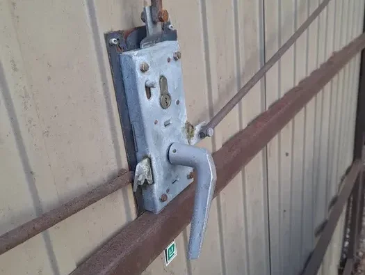 image Obraz na stronie internetowej przedstawia ręcznie wykonany mechanizm blokujący umieszczony na falistych metalowych drzwiach. Składa się on ze skomplikowanego montażu odsłoniętych kół zębatych i prętów, wraz z solidnym metalowym uchwytem, co jest przykładem rzemieślniczego kunsztu dostosowanego do napraw bram i okiennic w Warszawie.