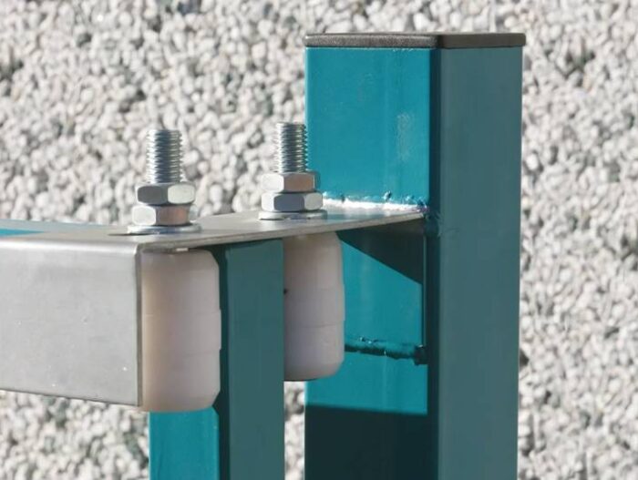 pol_pl_Rolka-prowadzaca-IBFM-O42-x-40-mm-nylonowa-ze-sruba-M14-typ-460-1013_4 Obraz na stronie internetowej przedstawia szczegółowy widok mechanizmu bramy przemysłowej, składającego się z połączenia elementów metalowych i plastikowych zabezpieczonych śrubami. Rama jest pomalowana na uderzający turkusowy kolor, wyróżniający się na tle teksturowanego szarego tła, podkreślając misterne rzemiosło związane z usługami naprawy i konserwacji bram oferowanymi w Warszawie.