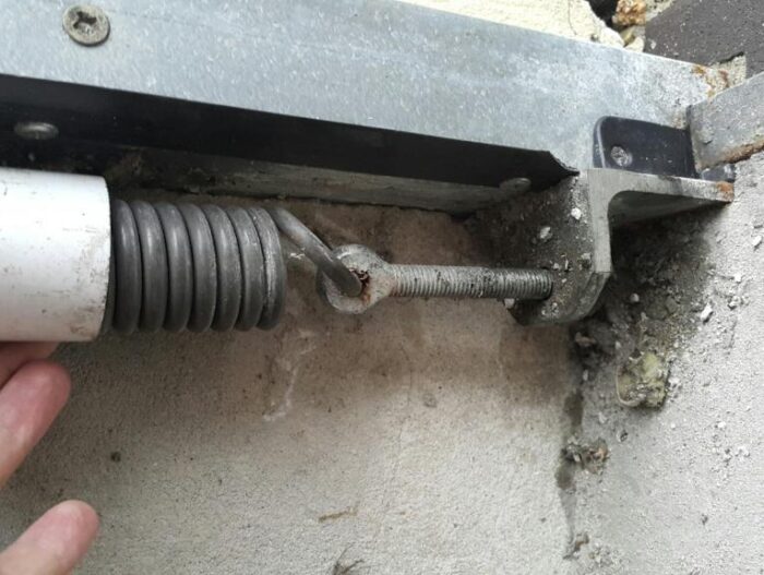 post-132897-0-72178100-1477487624 Obraz na stronie naprawy bramy warszawskiej, rolety i bariery pokazuje rękę dostrajającą metalową sprężynę naciągową połączoną z mechanizmem przesuwnym przymocowanym do teksturowanego betonowego tła. Sprężyna jest zabezpieczona wspornikiem, co ujawnia ślady zużycia konstrukcji.