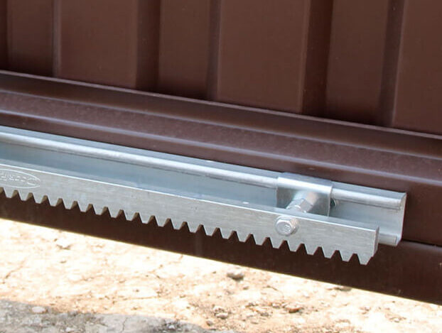 pravila-vyibora-zubchatoy-reyki-dlya-otkatnyih-vorot Obraz na stronie internetowej przedstawia widok z bliska metalowego mechanizmu bramy przesuwnej, ukazując jego misterny ząbkowany projekt toru. Tor jest bezpiecznie przymocowany do brązowej bramy, która jest lekko podniesiona nad żwirową powierzchnią, podkreślając szczegóły i kunszt związany z usługami naprawy bram w Warszawie.