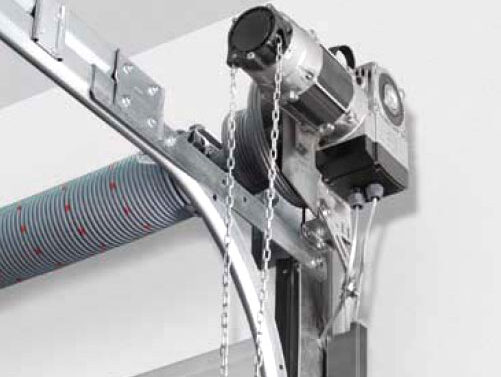 privod-sekcionnyh-vorot-ap26-big Na zdjęciu widać szczegółowy widok systemu otwierania drzwi garażowych z napędem silnikowym, zainstalowanego na suficie. Najbardziej rzuca się w oczy metalowy łańcuch przeplatany zakrzywioną szyną, która płynnie łączy się z mechanizmem sprężyny śrubowej. Silnik, bezpiecznie zamknięty, jest umieszczony nad szyną drzwi, podkreślając skomplikowaną konstrukcję i funkcjonalność typową dla wysokiej jakości usług naprawy i instalacji bram w Warszawie.