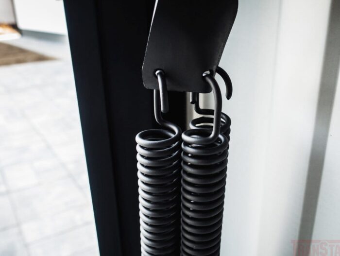 sprezyny-czarny-podklad-2 Na zdjęciu widać z bliska dwie pionowo wiszące czarne metalowe sprężyny, bezpiecznie przymocowane do haków na metalowym uchwycie. Tło przedstawia delikatnie rozmytą podłogę, podkreślając sprężyny, często kluczowe w procesach naprawy bram, rolet i barier w Warszawie.