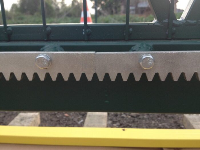 vidy-zubchatoj-rejki-dlya-otkatnyh-vorot Obraz na stronie internetowej poświęconej naprawom bram, rolet i barier w Warszawie przedstawia szczegółowy widok metalowego mechanizmu zębatego z ząbkowaną krawędzią. Na pierwszym planie wyraźnie widać dwa widoczne rygle na górze mechanizmu. Żółta szyna rozciąga się poziomo przez dolną część ramy, a tło jest delikatnie rozmyte, aby skupić uwagę na elementach mechanicznych.